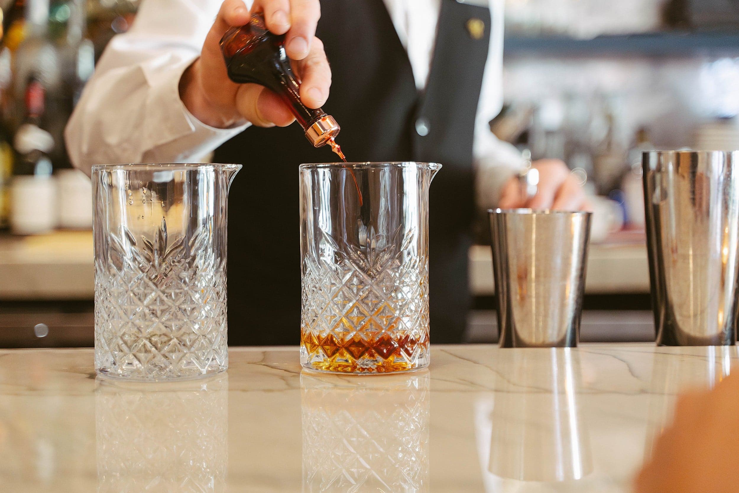 Bartender adds bitters from a dropper bottle into a crystal mixing glass on a marble bar.