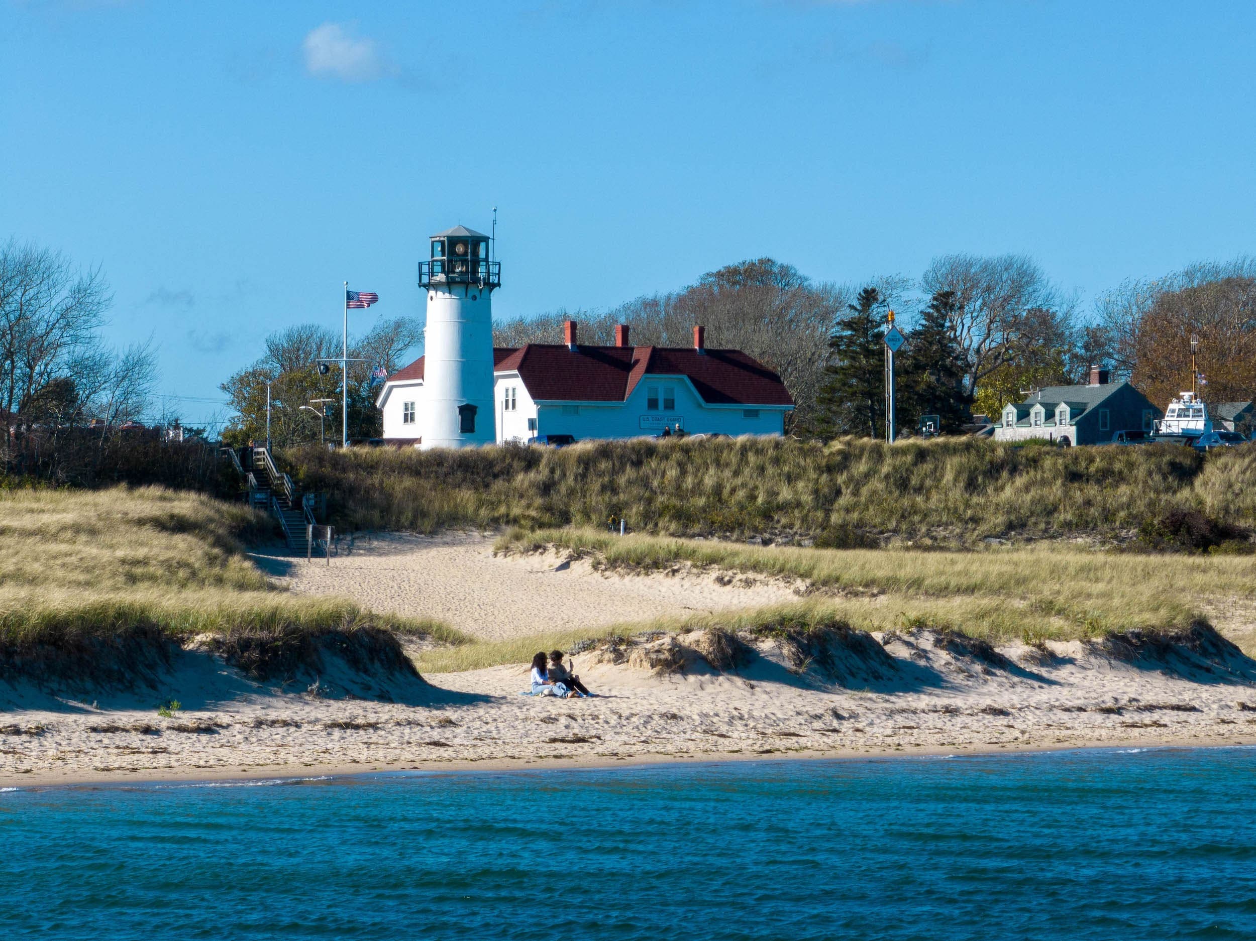 A couple sitting on the beach with a lighthouse and beach houses behind them
