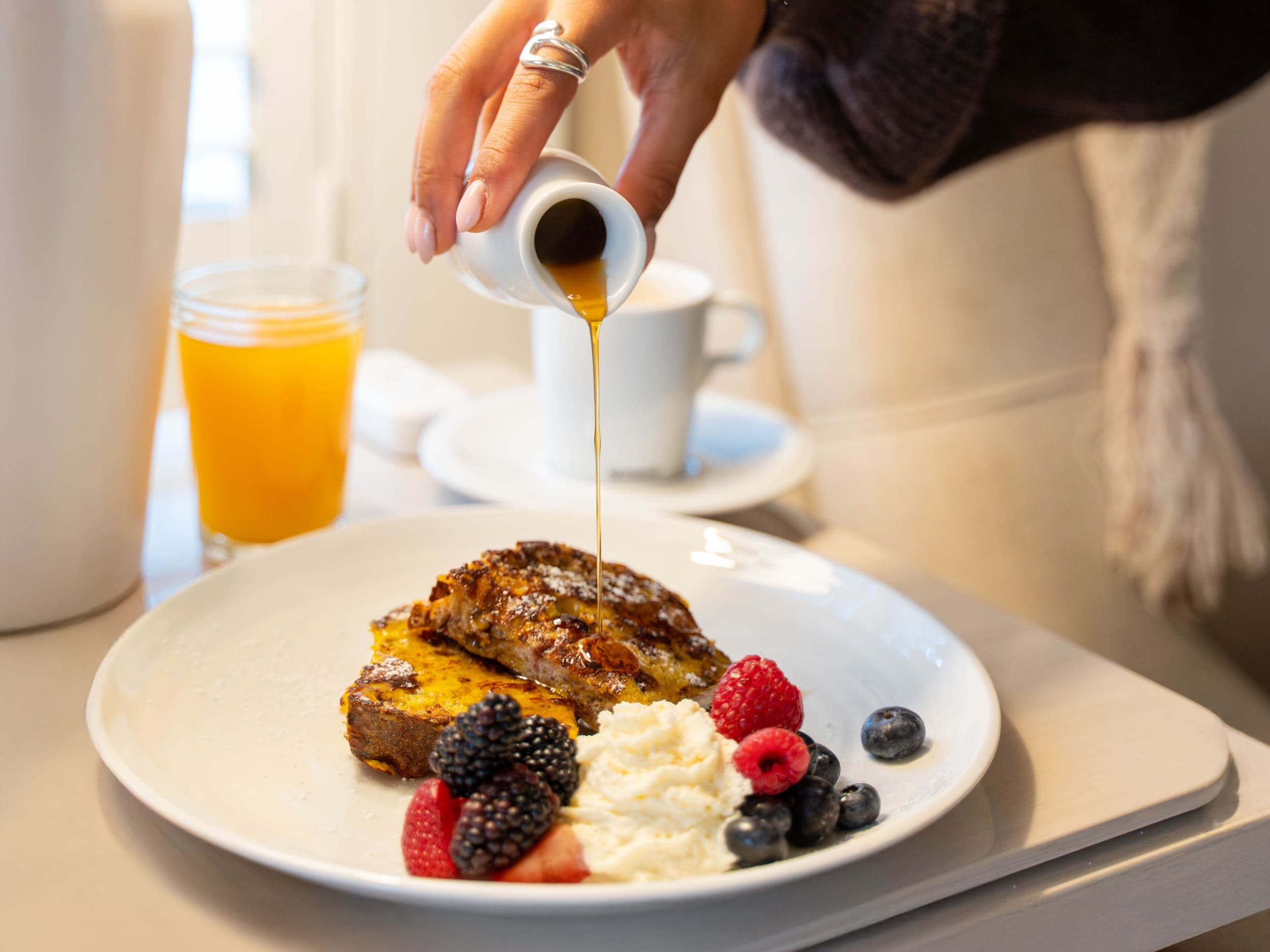 A woman's hand pouring syrup over french toast with berries and whipped cream